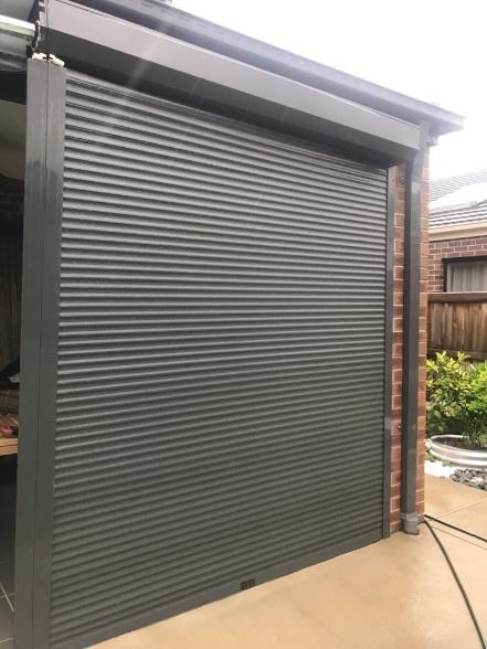 Charcoal security roller shutter fully closed over a residential patio opening beside an exterior brick wall.