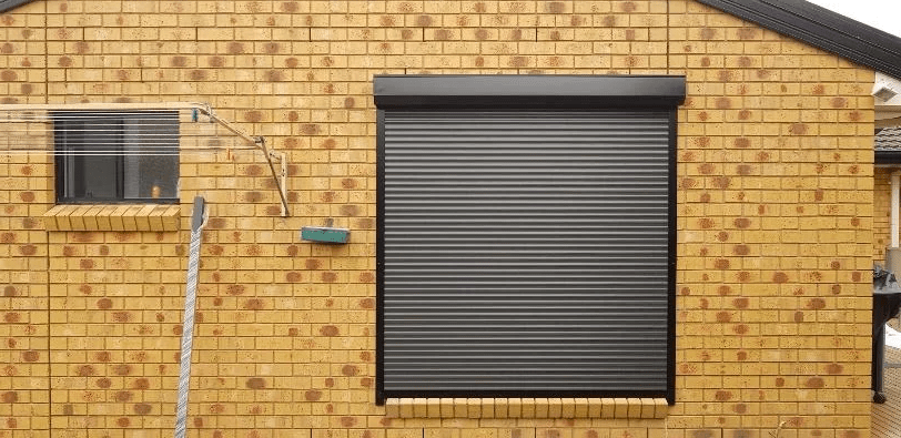 Charcoal security roller shutter fully closed over a residential window on an exterior yellow brick wall.